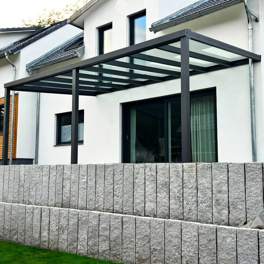Anthrazitfarbene Terrassenüberdachung mit Glasdach, harmonisch integriert in eine Terrasse mit Steinpalisaden in Langenfeld / Bergisch Gladbach