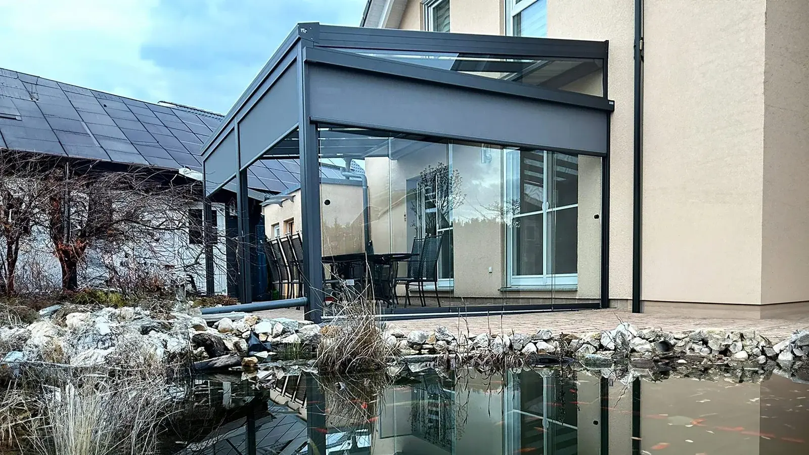 Moderner Wintergarten aus Glas mit Anthrazit-Profilen und Schiebesystemen neben einem Gartenteich in Bergisch Gladbach